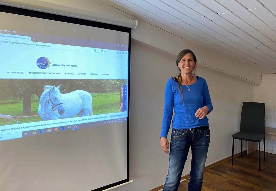 Jeanette Karlen - steht lachend neben einer Leinwand und hält einen Vortrag.